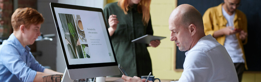 Cyber-Versicherung Eine Gruppe von Personen arbeitet gemeinsam in einem modernen Büro. Im Vordergrund sitzt ein Mann vor einem Computerbildschirm, der eine Website zeigt. Im Hintergrund stehen zwei Personen, die ein Tablet und ein Smartphone benutzen. Ein weiterer Mann sitzt seitlich im Bild an einem Tisch.